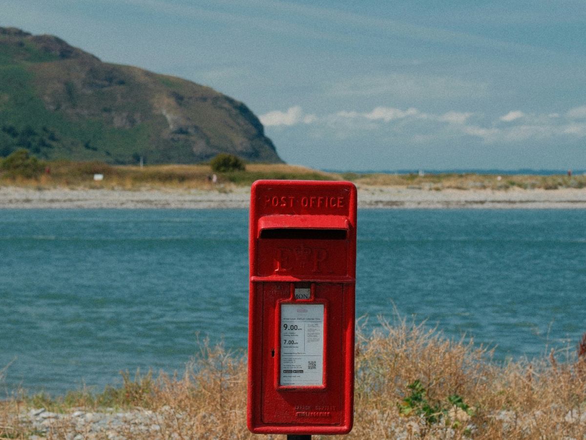 A Chicago manufacturing manager auditing a batch of parts invoices and applying forever stamps from a roll of 100 with an industrial production floor in the background.